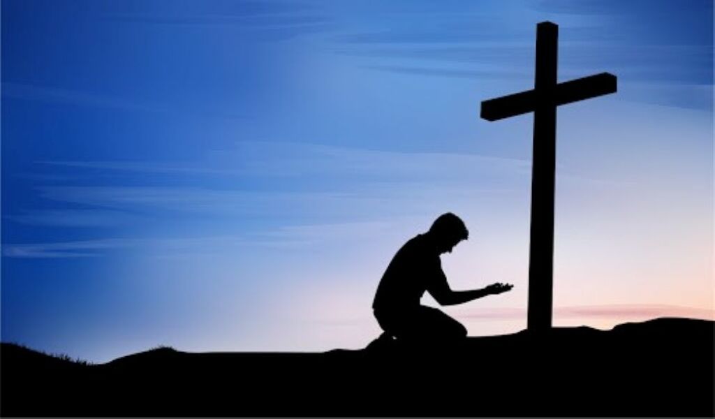 Silhouette of a kneeling person beside a cross at sunset on a hill