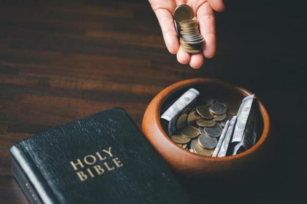 Hand placing coins into a bowl beside a Holy Bible on a wooden table