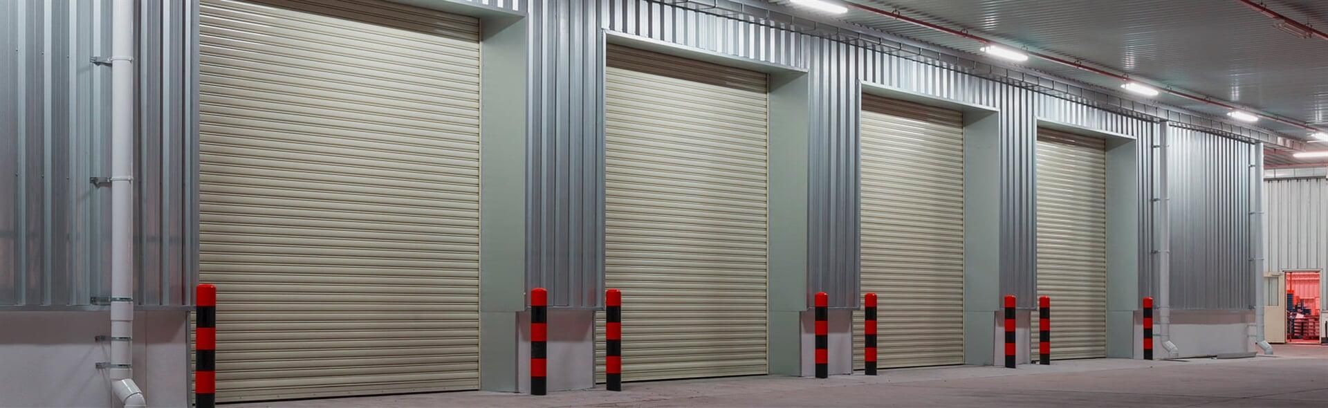 Gran almacén industrial gris con puertas enrollables cerradas y bolardos de seguridad rojos.
