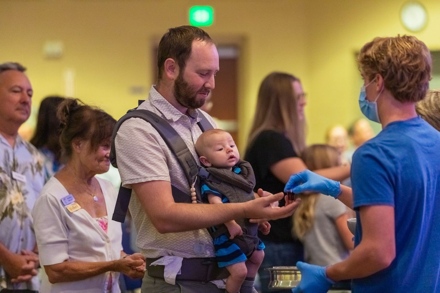 A man is holding a baby in a carrier while talking to a man in a blue shirt.