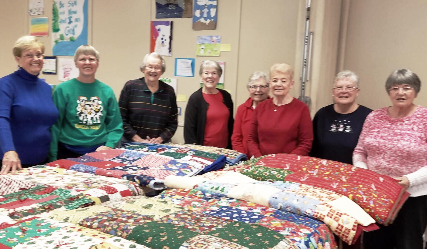 A group of women are standing around a table with quilts on it.