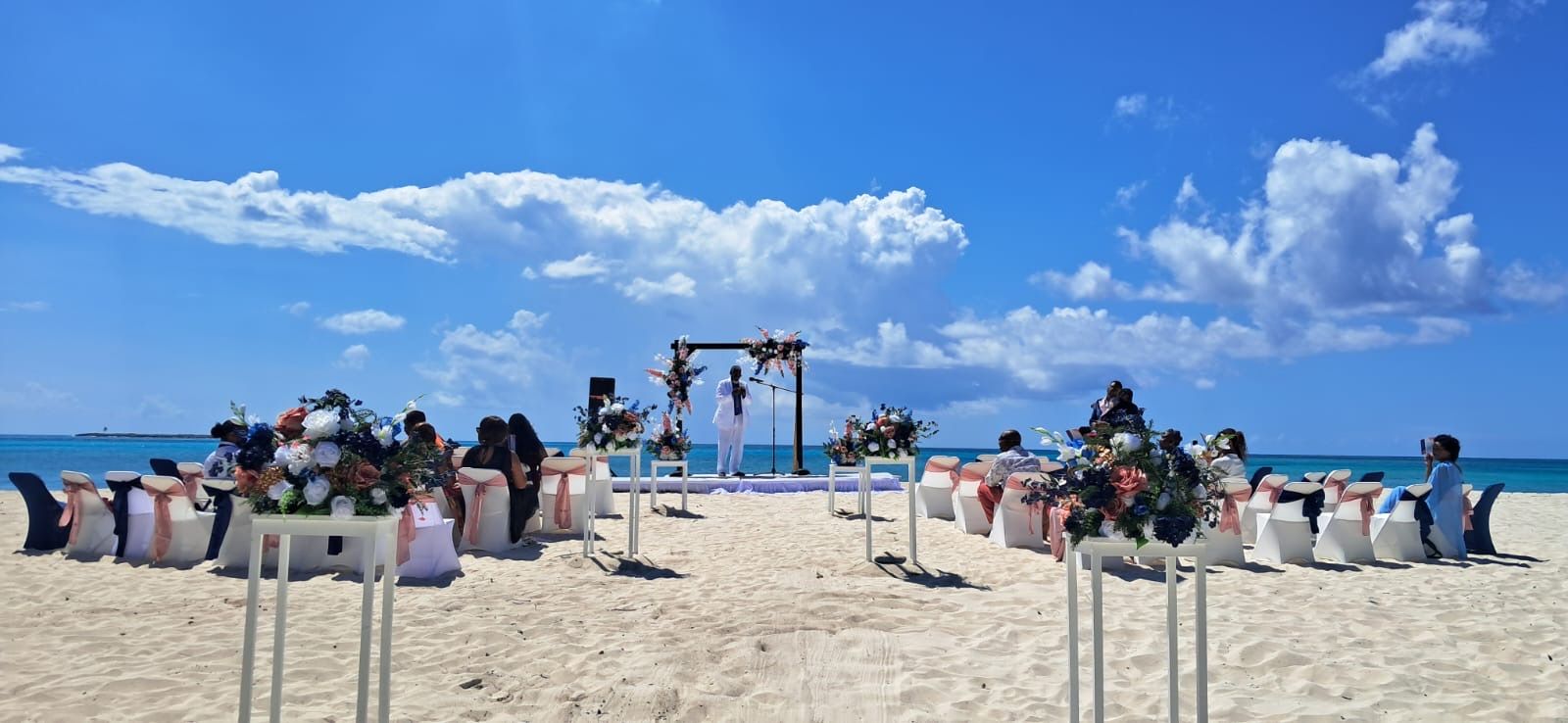 Beach wedding setup with chairs and floral arrangements, blue sky, and ocean background.