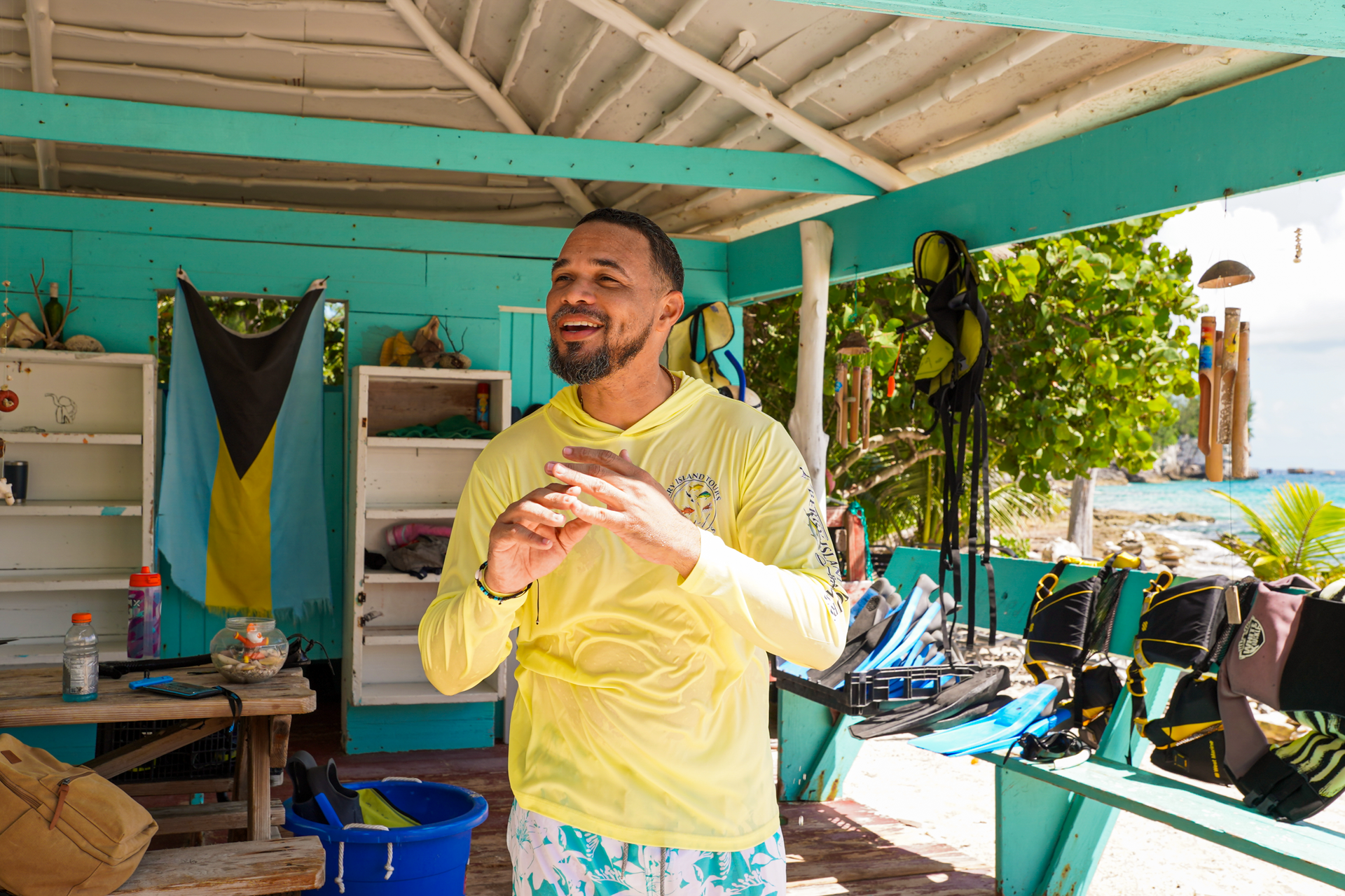 Man in yellow shirt smiles, adjusting his watch; turquoise building, ocean, and equipment visible.