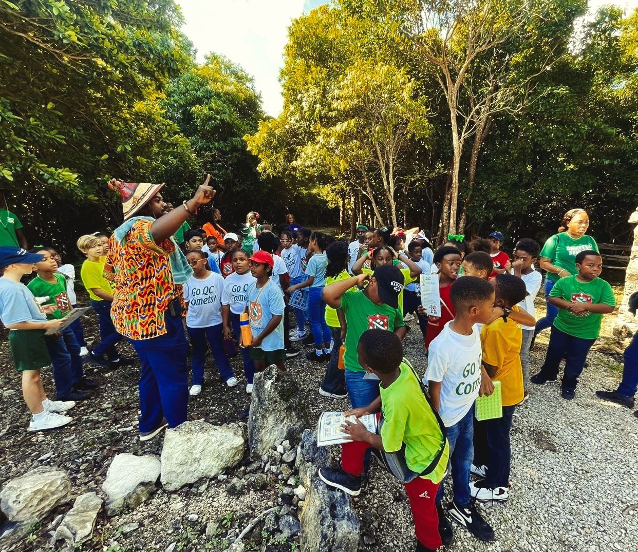 Group of children listening to a person pointing in a wooded area. The children wear various colors.
