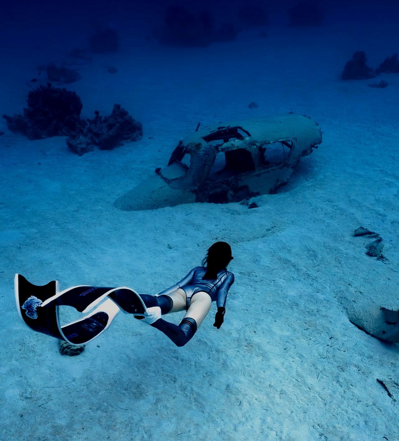 Freediver swims past submerged airplane wreckage on sandy ocean floor.