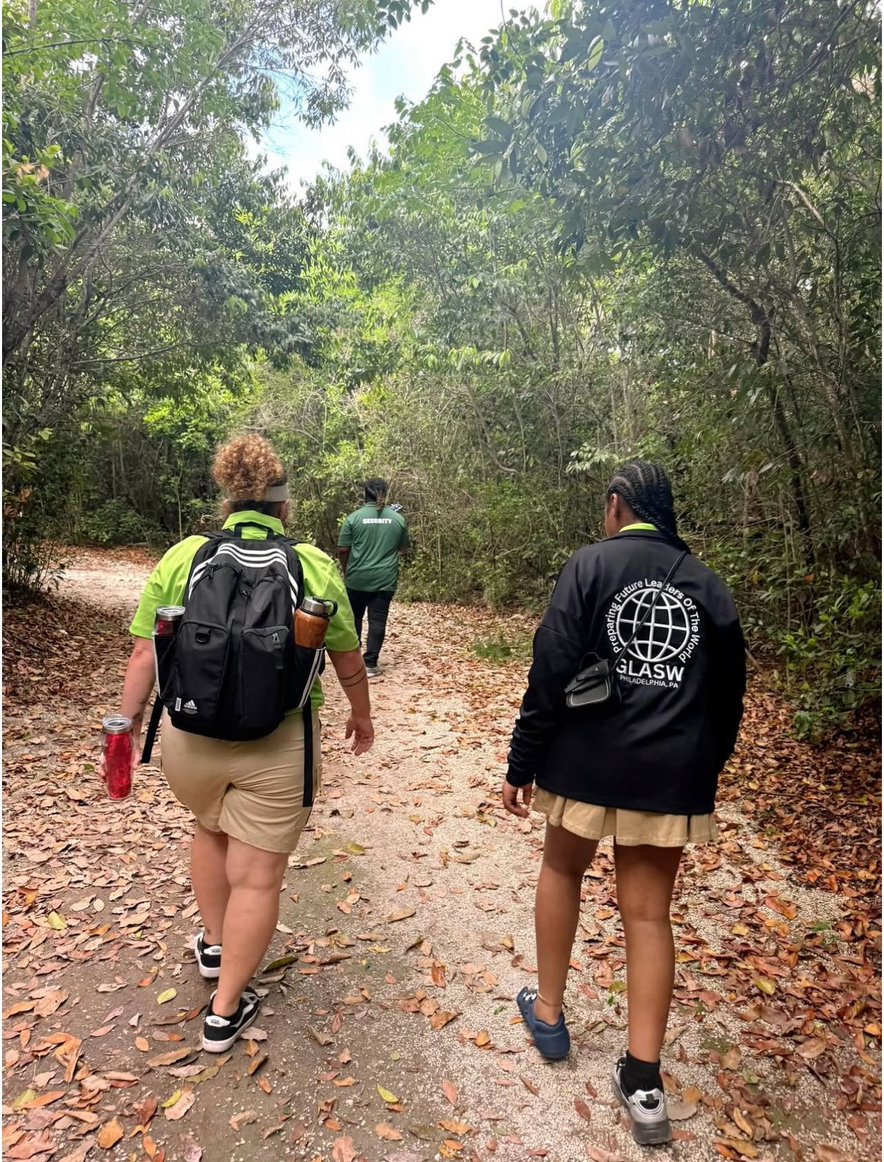 Three people walk on a leaf-covered path in a wooded area. One carries a backpack and water bottle.