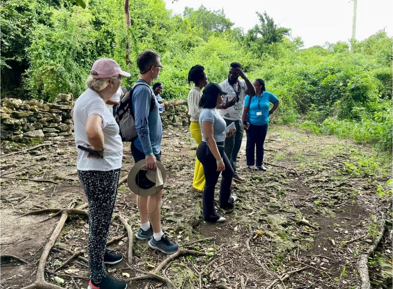 Group of people on a dirt path in a wooded area, listening to a person wearing a blue shirt.