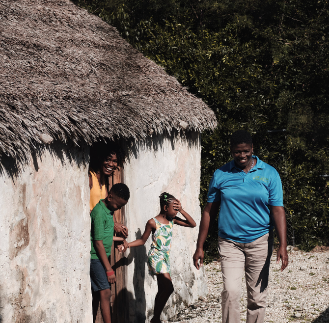 Family outside a thatched-roof structure. Person in blue shirt walks toward camera. Others look out from the doorway.