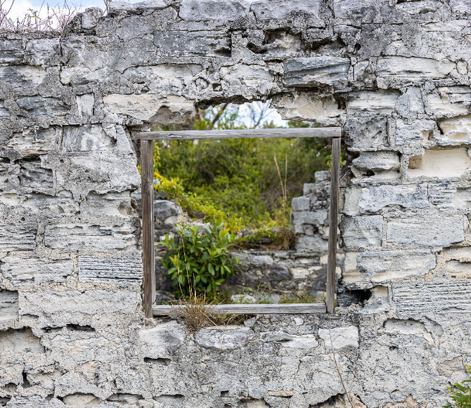 Ruined stone wall with wooden window frame; view of green foliage through the opening.
