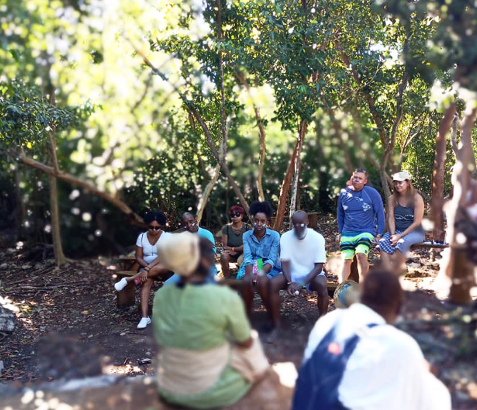 Group sitting on benches in a wooded area, listening to someone. Sunlight filters through the trees.