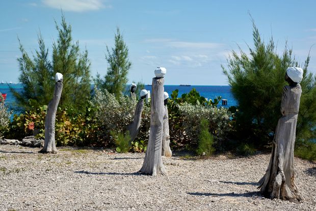 Wooden sculptures topped with white spheres stand on a pebble ground, with the ocean in the background.