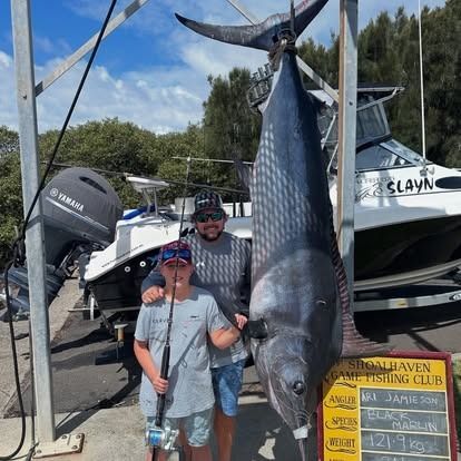 Man and boy pose with a large black marlin suspended at a dock.