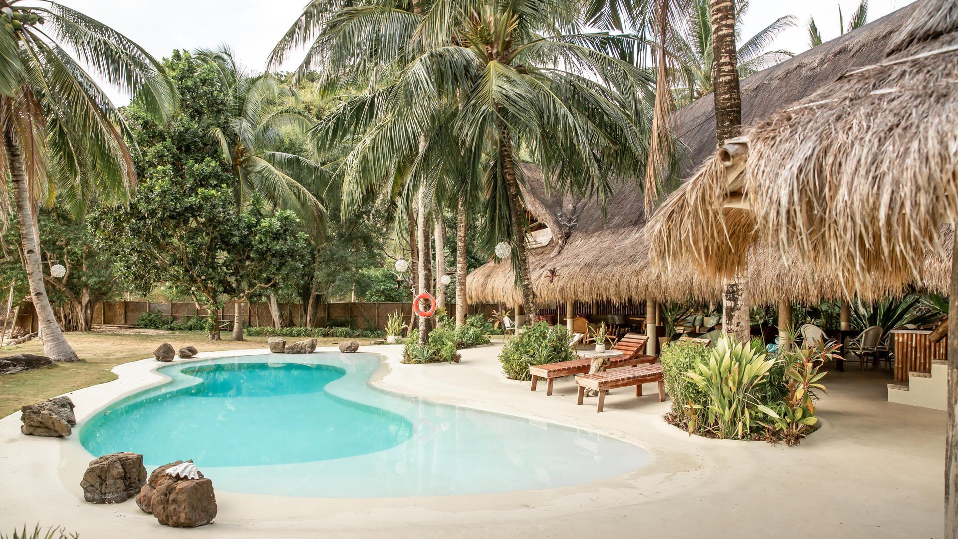A large swimming pool surrounded by palm trees and thatched huts.