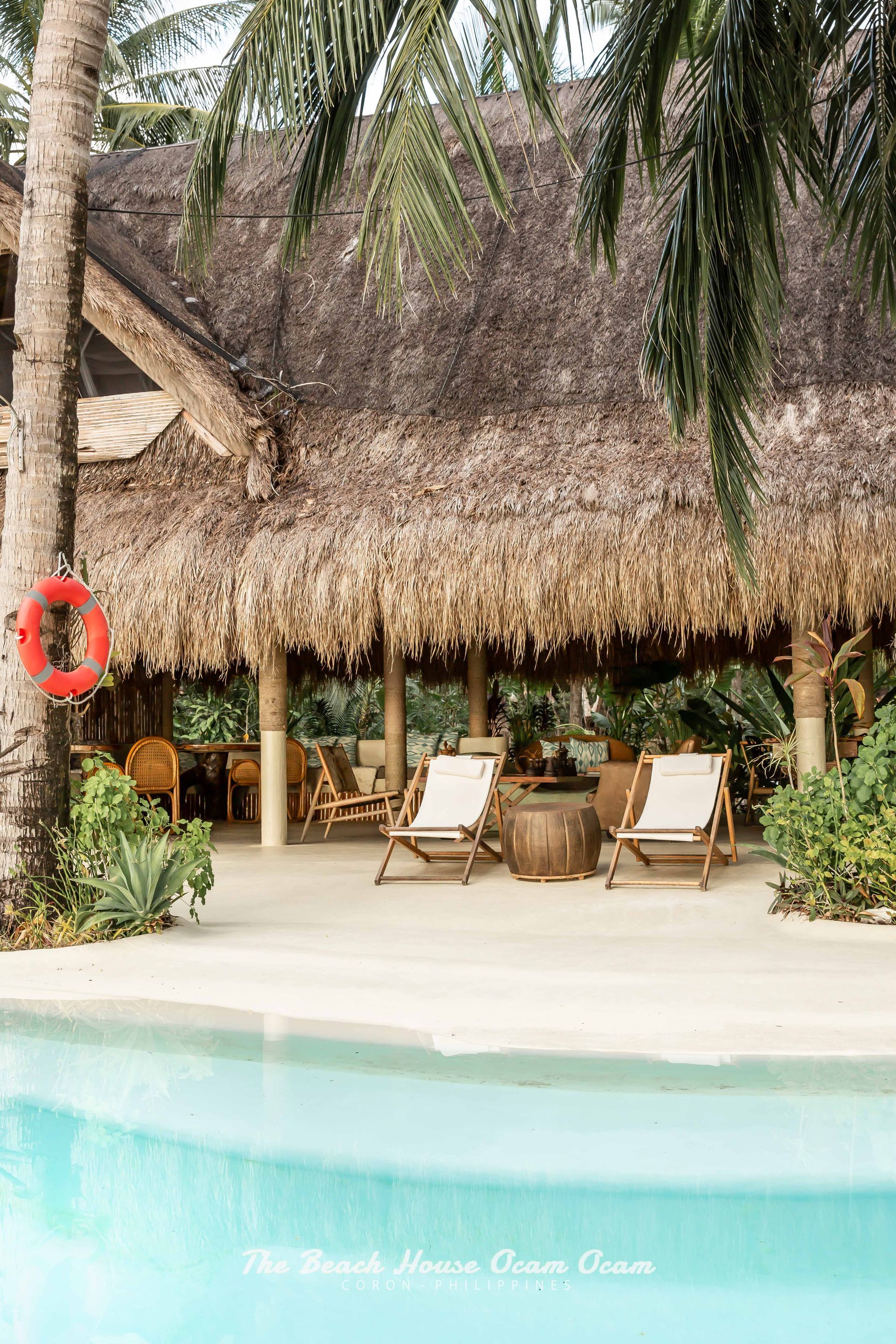 A swimming pool surrounded by chairs and palm trees with a thatched roof.