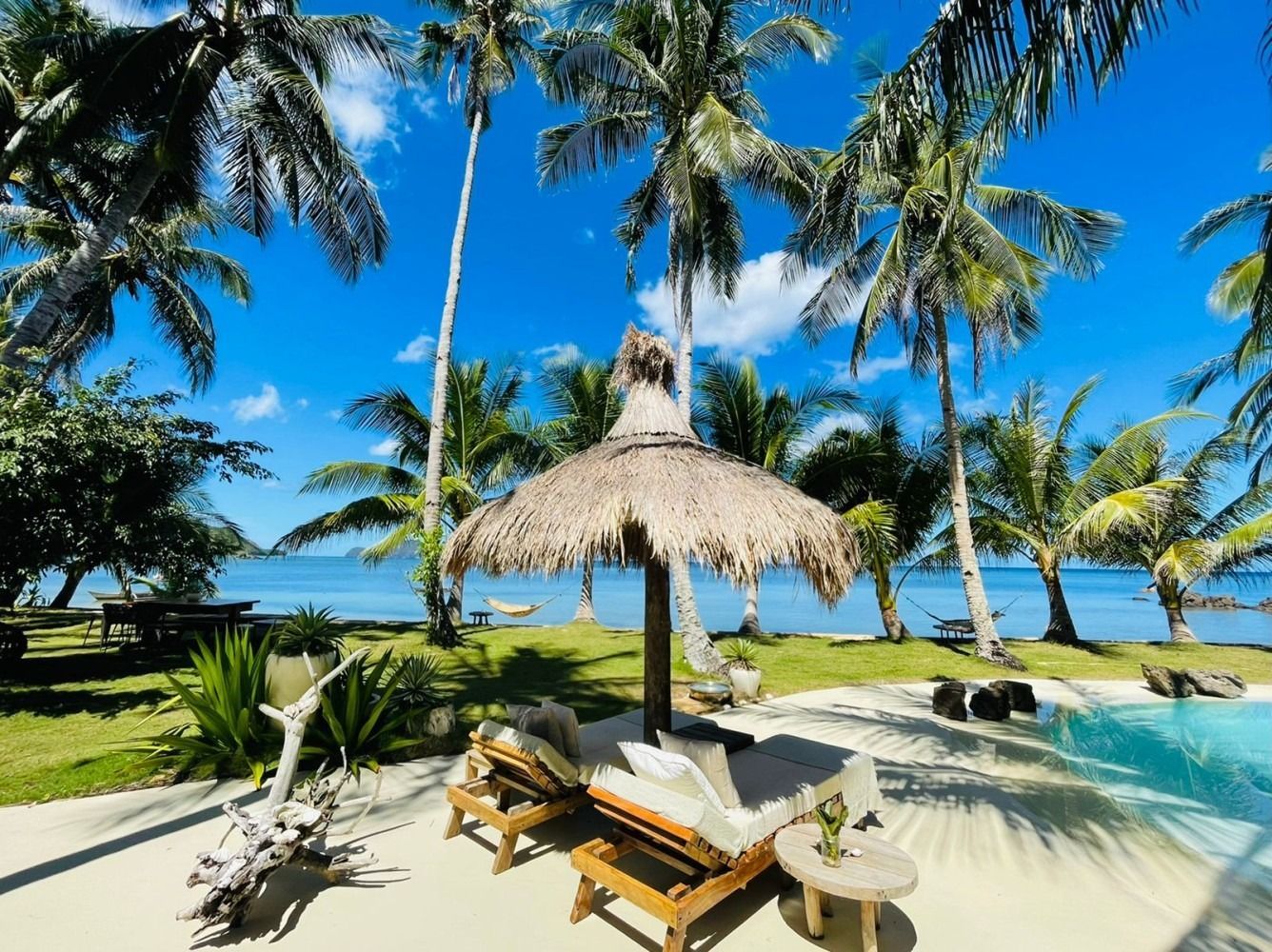 A thatched umbrella sits on a sandy beach next to a swimming pool surrounded by palm trees.