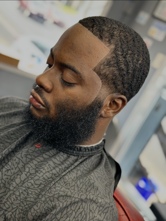 Man with a groomed beard and wavy haircut, wearing a barber's cape, eyes closed.