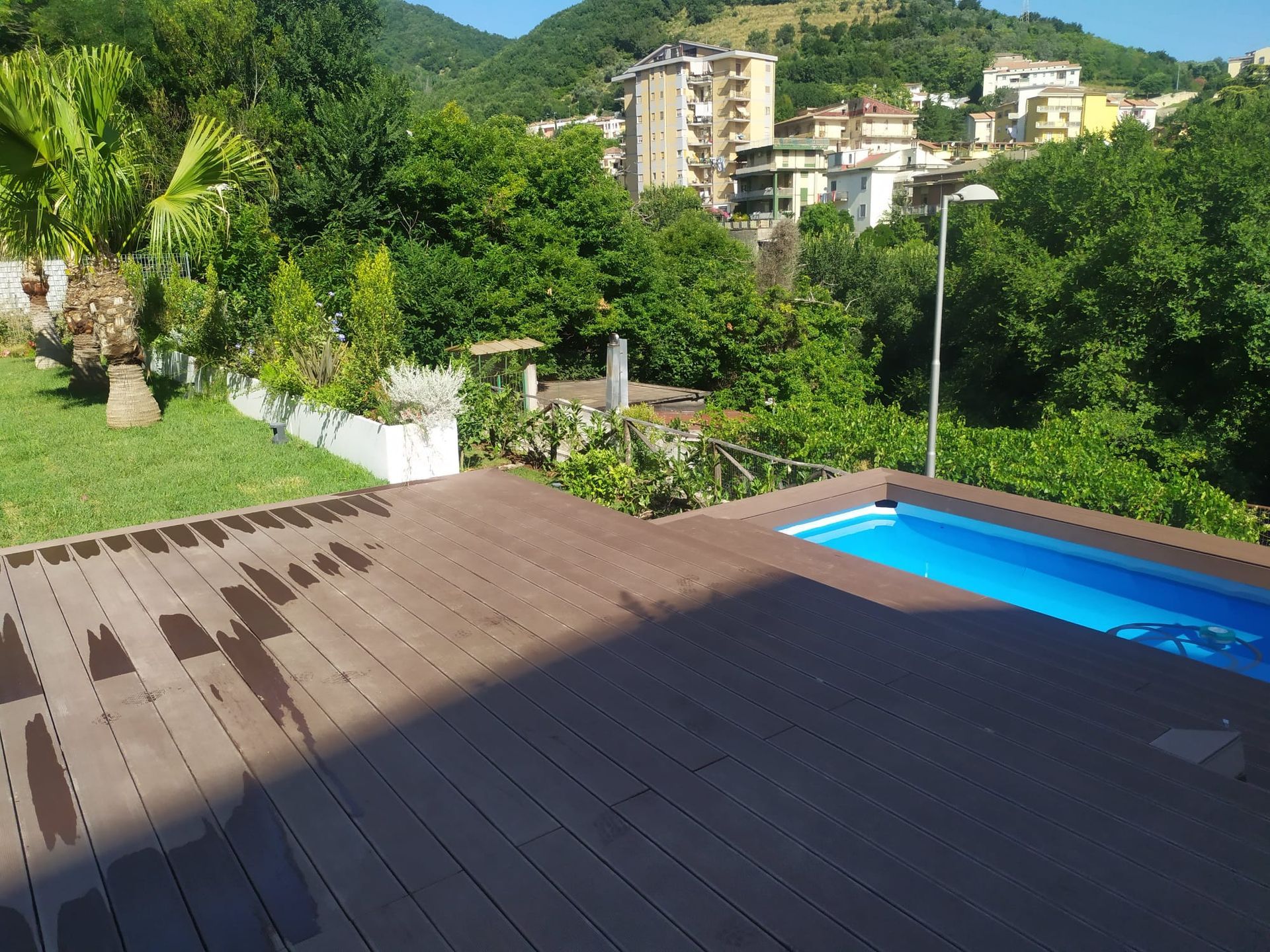 Un ponte di legno circonda una piccola piscina blu, circondata da una vegetazione lussureggiante e con una città sullo sfondo.