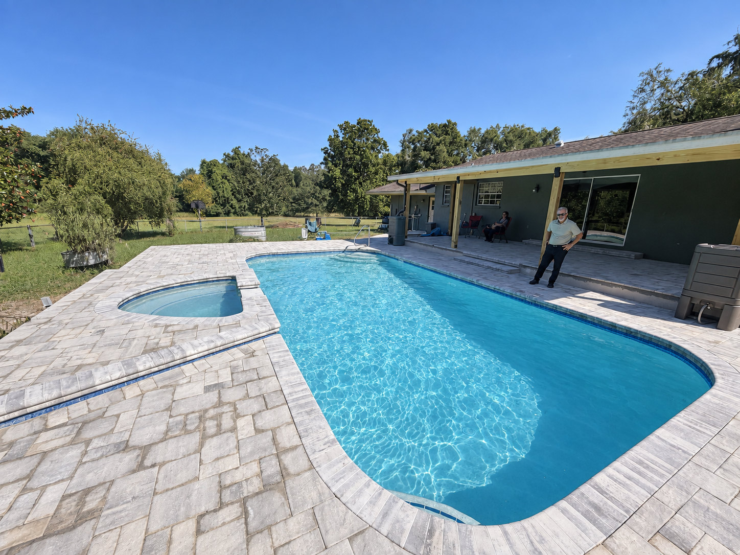 Backyard patio with a curved swimming pool, stone deck, and a person standing by the covered porch
