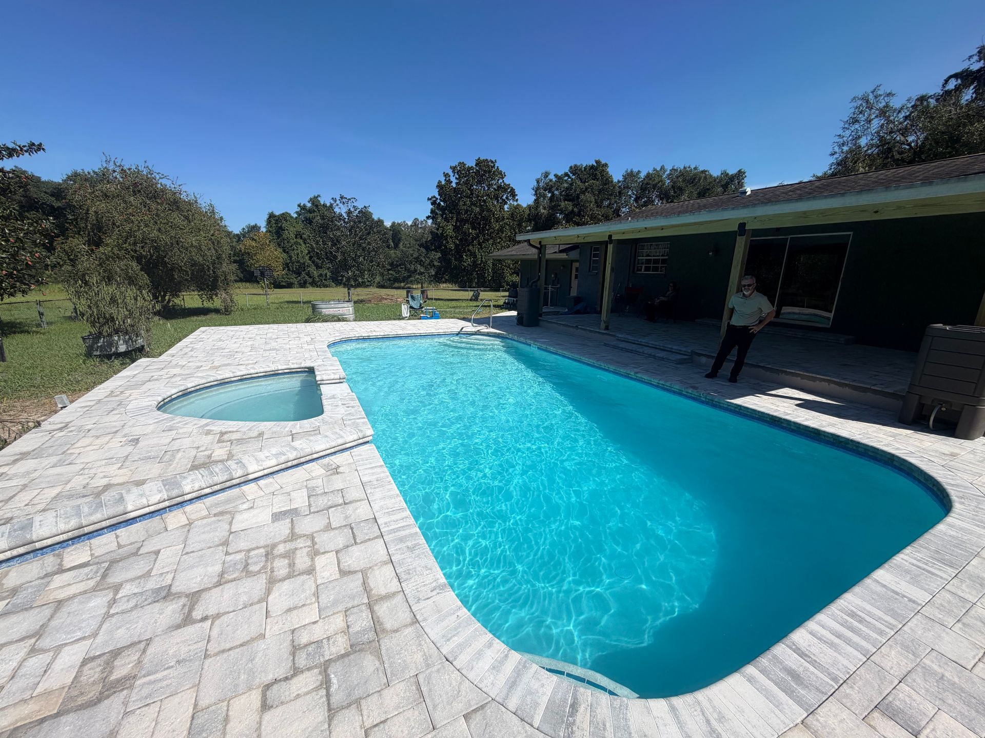 Backyard pool with bright blue water, stone patio, and shaded patio seating area on a sunny day