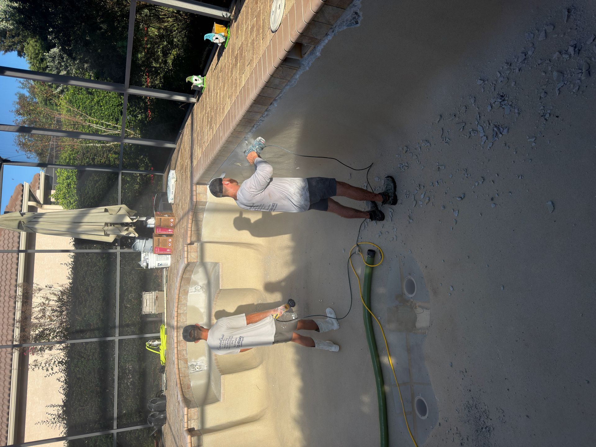 Two people in white shirts and shorts stand on a sunny tennis court near a fence.