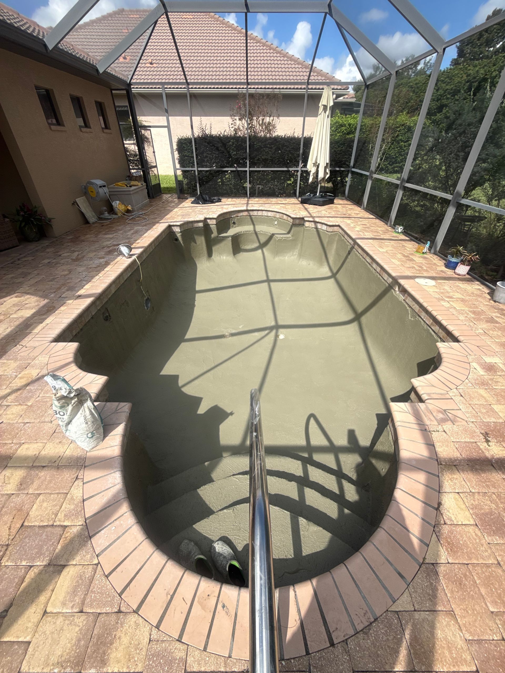 Covered backyard swimming pool with green water, seen under a screened patio and brick deck