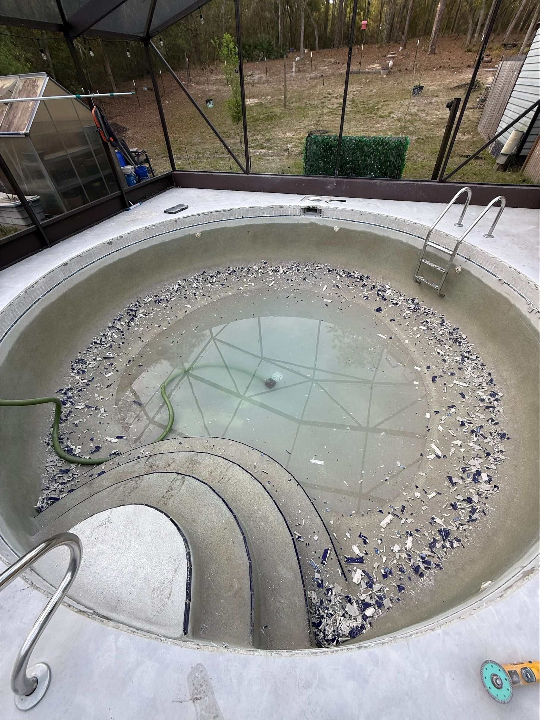 Empty round outdoor pool with gray steps, shallow water, and scattered debris in a fenced yard