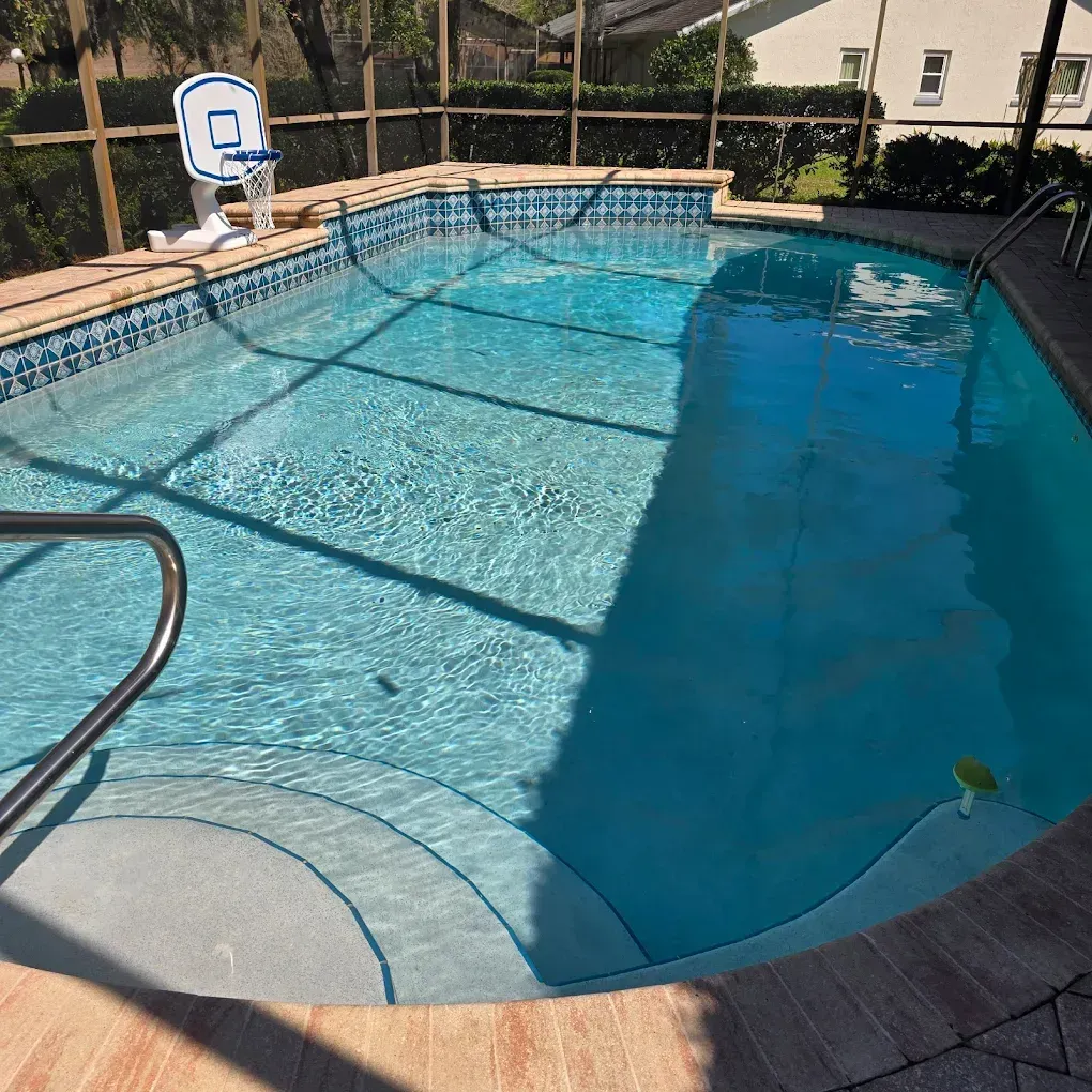 Outdoor backyard swimming pool with blue water, tiled deck, and metal handrails, viewed in bright sunlight