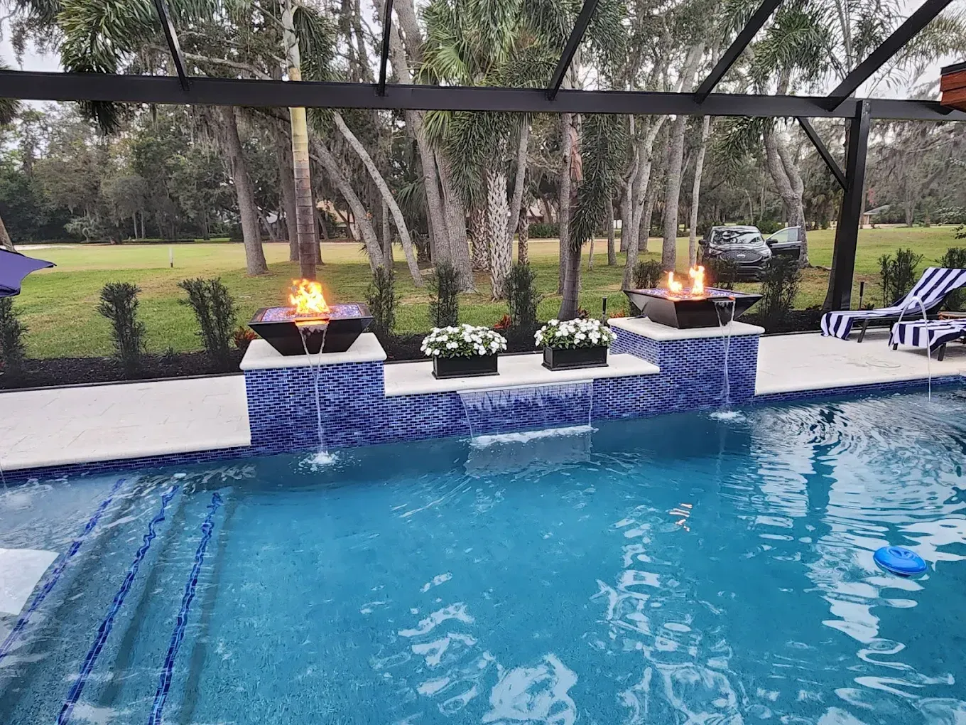 Backyard pool with blue water, tiled spa ledges, lit fire bowls, and lounge chairs under a screened enclosure