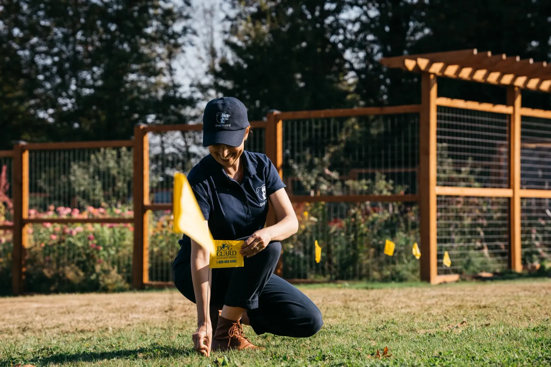 A person in a dark uniform and cap kneels in a grassy field, placing a small yellow marker into the ground.