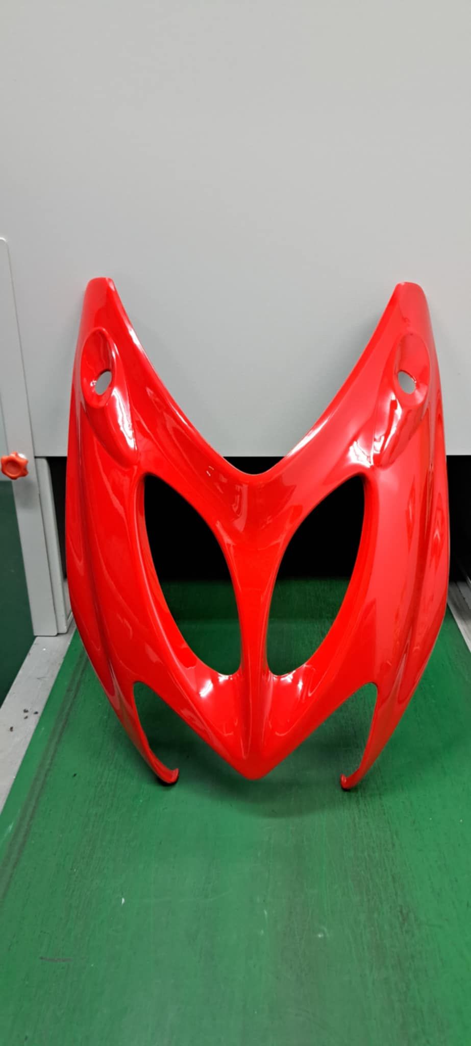 灰色牆面上，綠色表面上矗立著一個紅色光澤的抽象面具或頭盔狀雕塑。