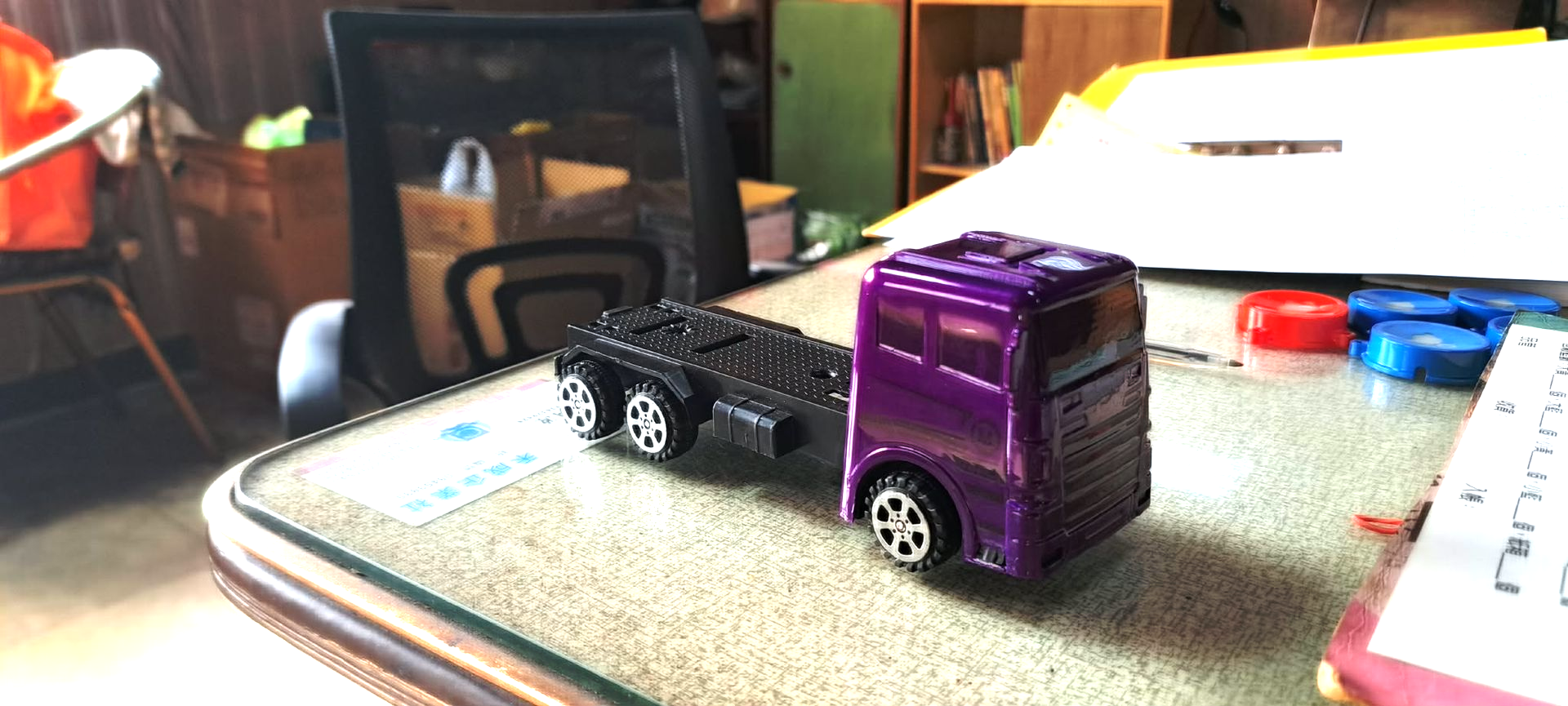 桌面上放著兩輛玩具卡車，一輛紫色，一輛黑色，背景裡有一輛玩具汽車。