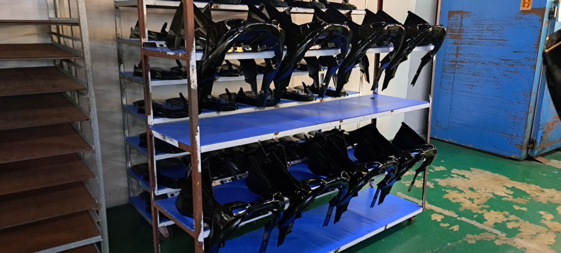 車間裡，白色架子上擺放著一排排黑色機械零件，架子下方是藍色的箱子，後面是一面藍色的牆。