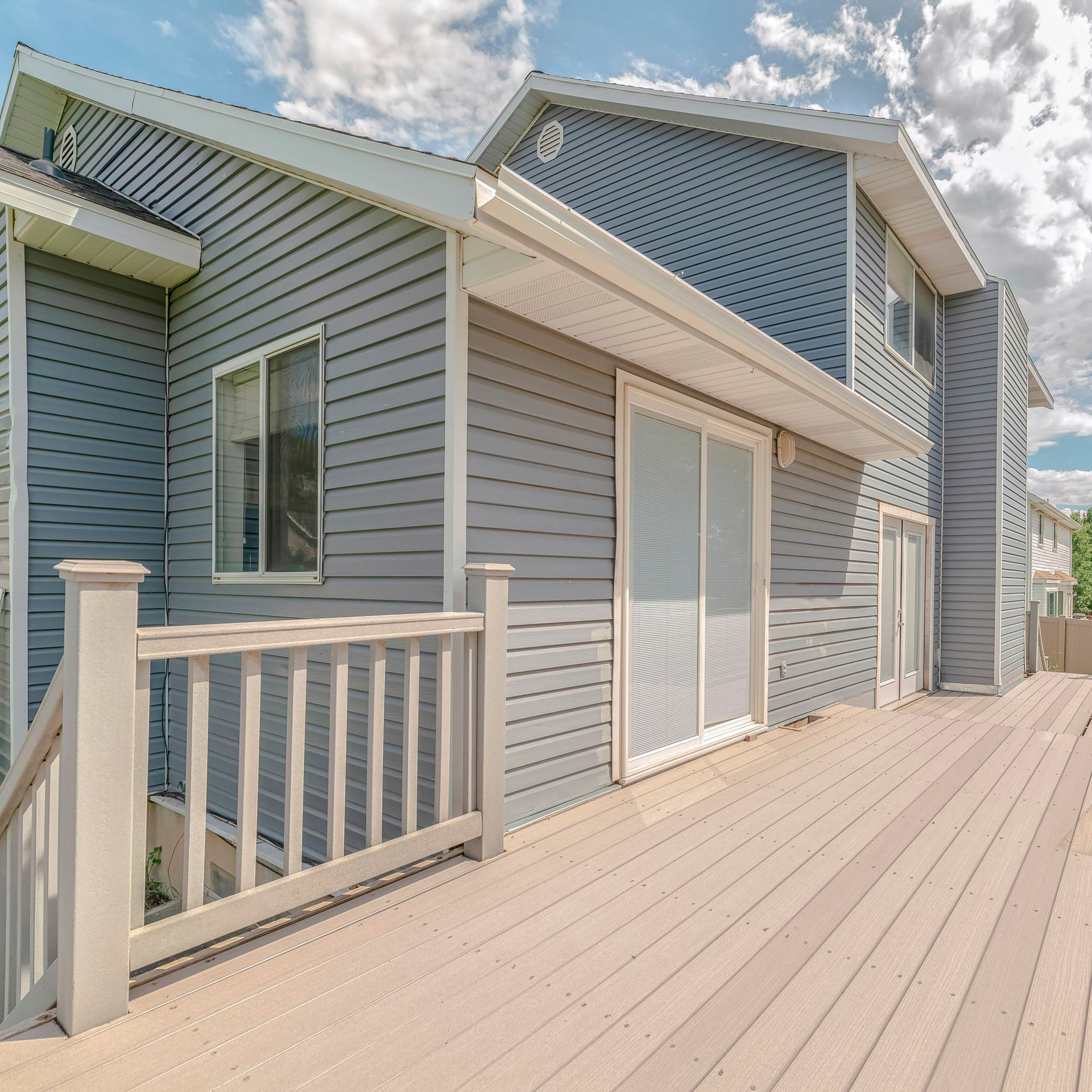 Blue-gray house with a wide wooden deck and sliding glass doors under a partly cloudy sky
