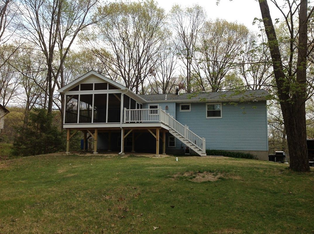 Blue house with screened porch and front stairs in a grassy yard, surrounded by tall trees