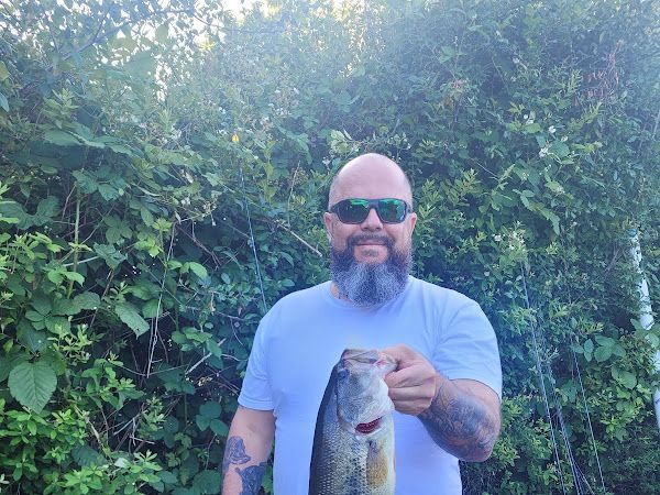 Man in sunglasses holds up a fish, standing in front of lush green foliage.