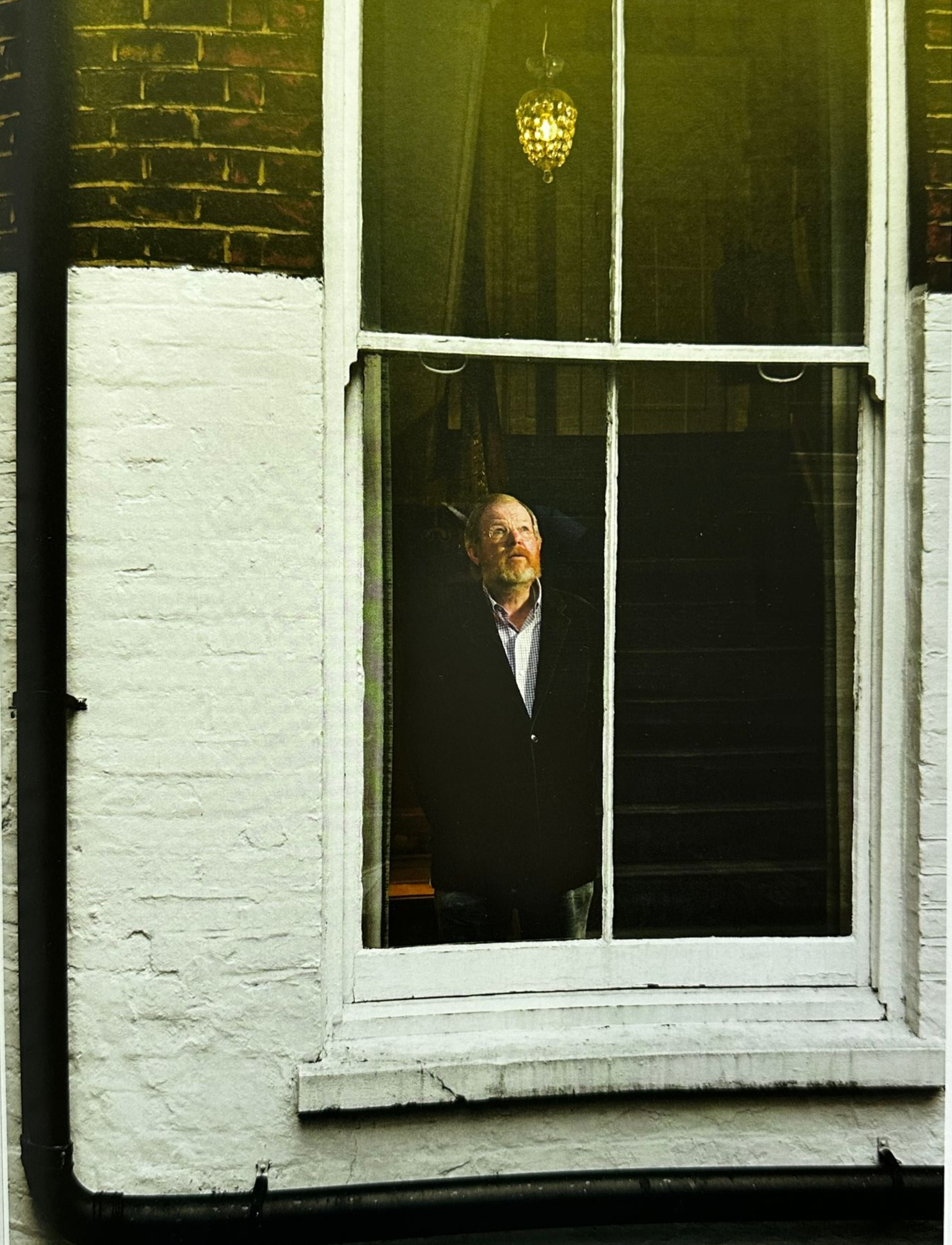 A man in a dark suit stands inside a window of a brick building.