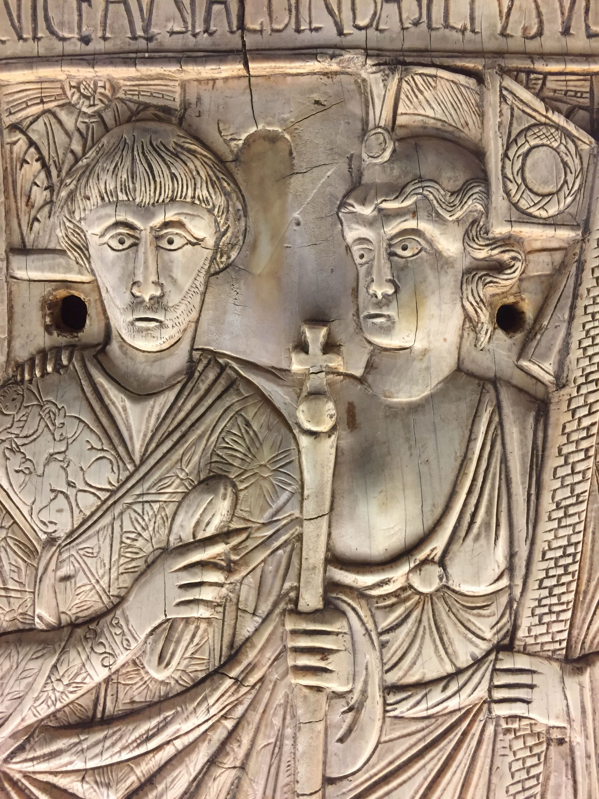 Ancient carved relief of two robed figures holding a staff or cross, with decorative geometric background