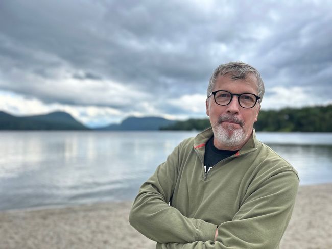 Man with glasses and gray beard standing by a lake under cloudy skies, arms crossed, wearing an olive jacket