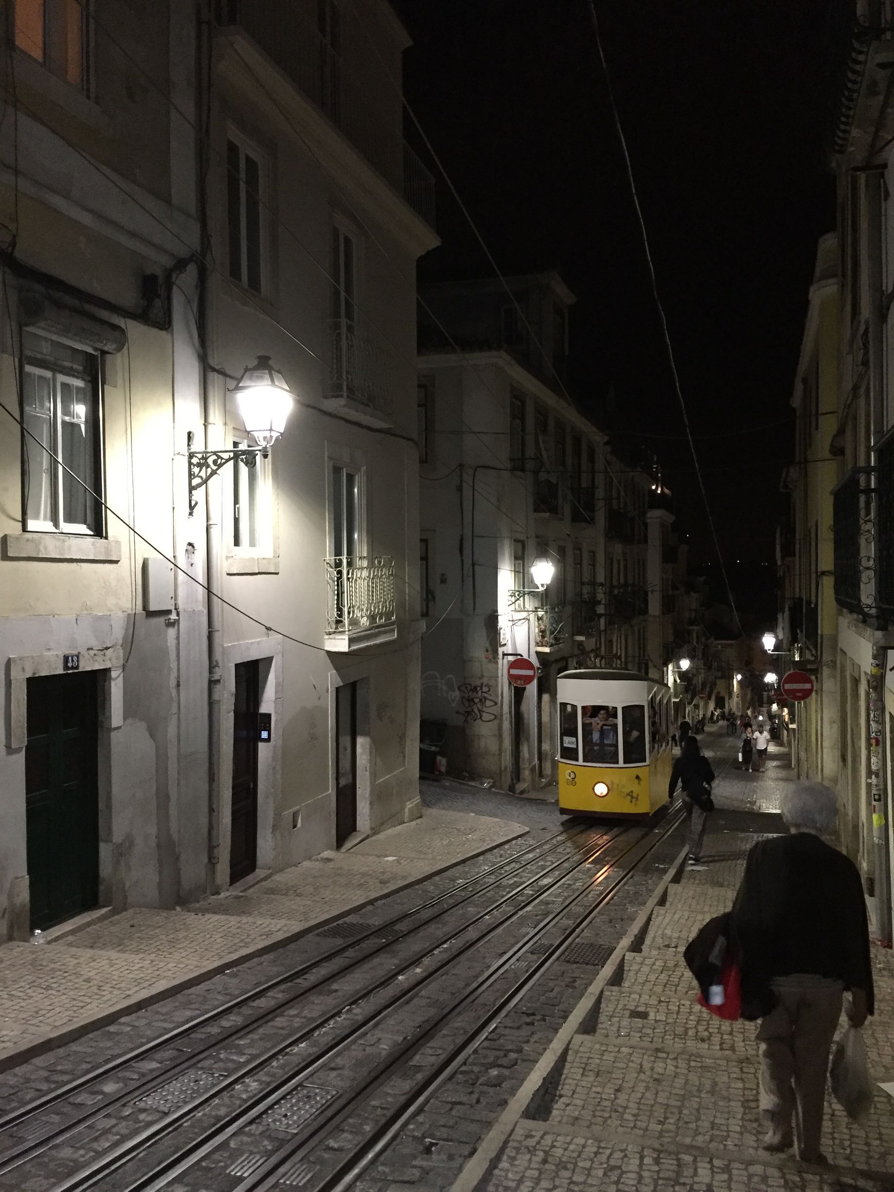 Nighttime street with tram tracks, a yellow tram, and pedestrians on a narrow cobblestone road.
