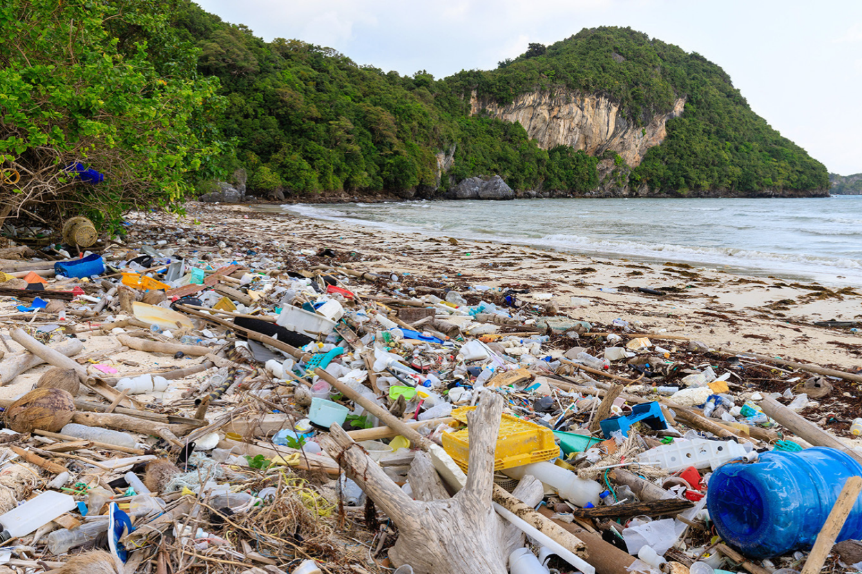 Trash-strewn beach with plastic debris along the shoreline beneath a green rocky hill