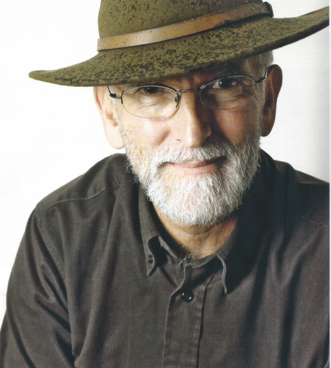 Portrait of a bearded man in glasses and a brown hat, wearing a dark shirt, against a plain background