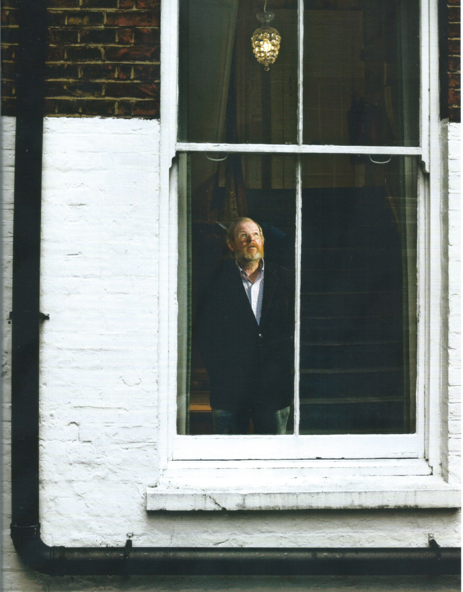 A man in a dark suit stands inside a window of a brick building.