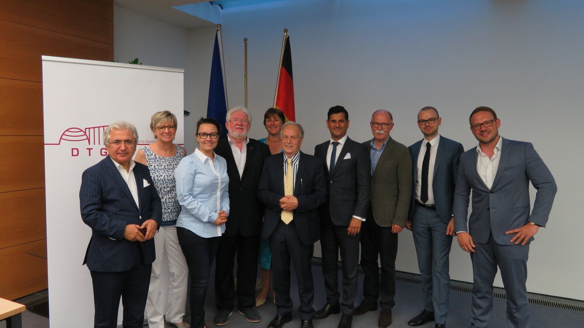 Eine Gruppe von Menschen posiert für ein Foto. Im Hintergrund sind die Flaggen der EU und Deutschlands zu sehen. Weißer Hintergrund mit Logo.