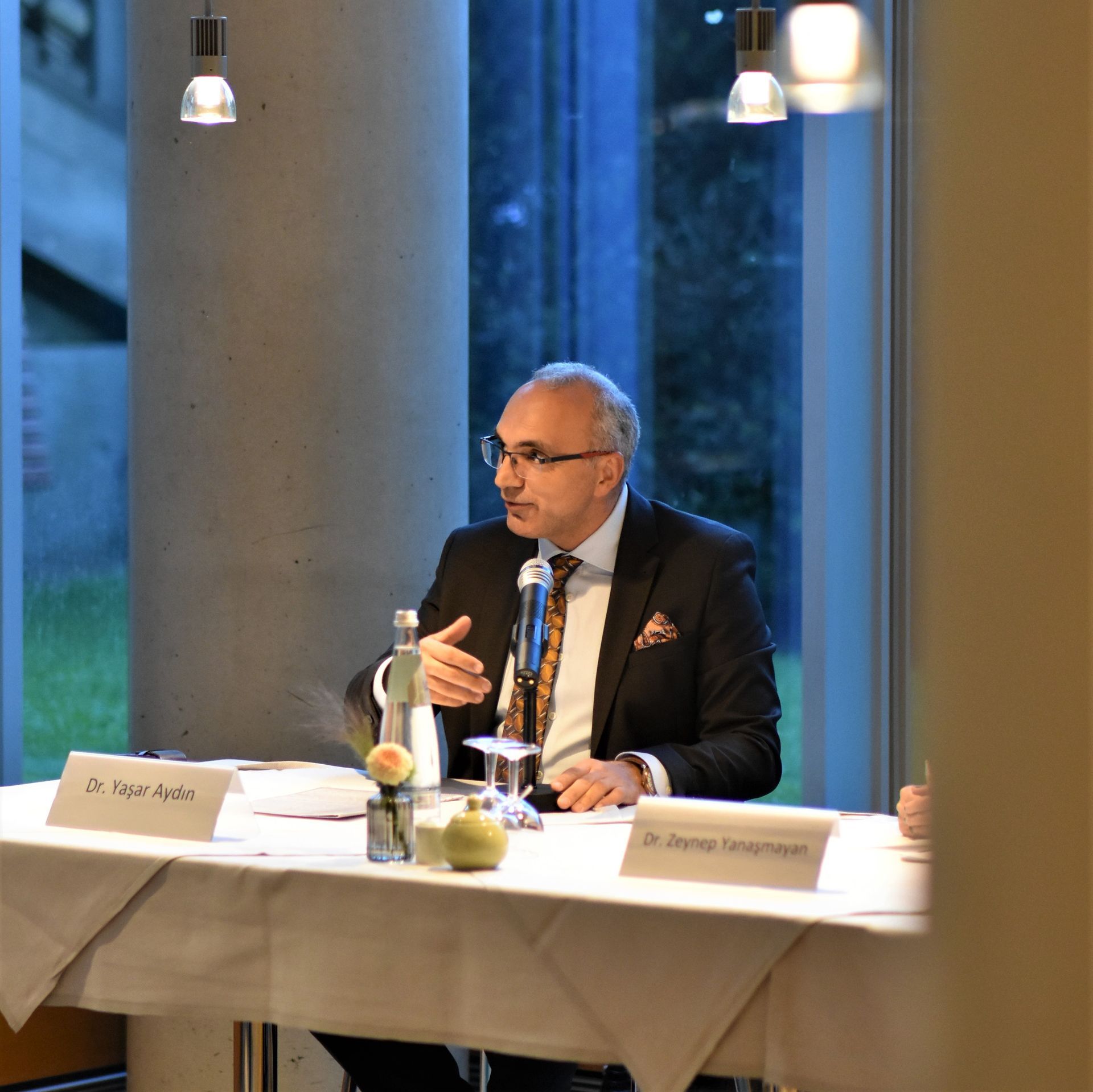 Ein Mann spricht an einem Tisch mit Mikrofon, Dokumenten und Wassergläsern in einer Konferenzsituation.