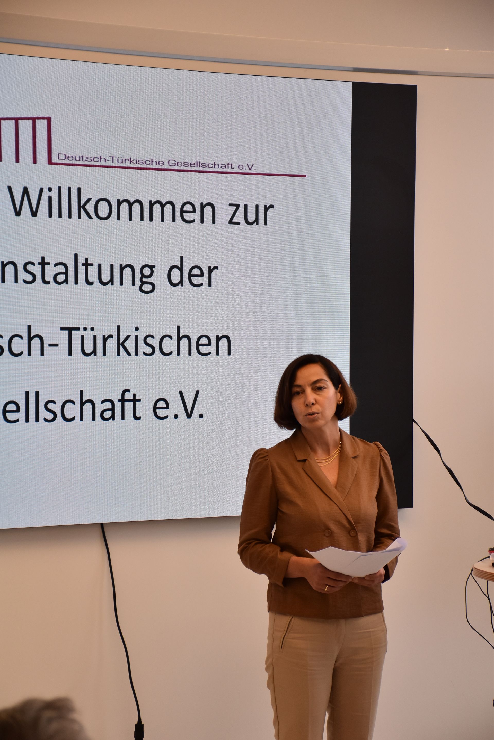 Eine Frau in einem braunen Blazer spricht bei einer Veranstaltung vor einer Leinwand mit deutschem Text.