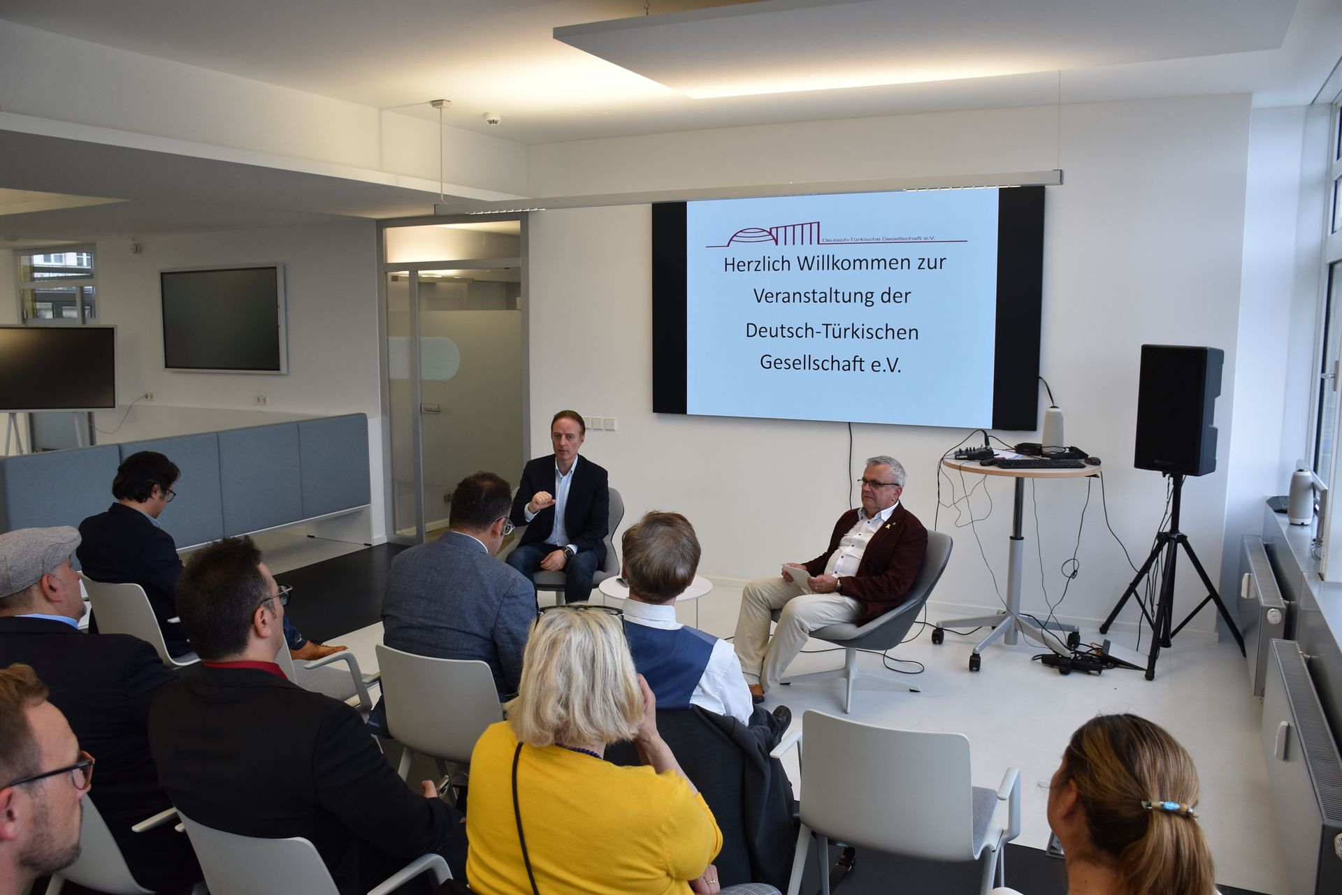 Eine Podiumsdiskussion in einem lichtdurchfluteten Raum mit zwei Männern auf der Bühne, einer Leinwand hinter ihnen und einem Publikum.