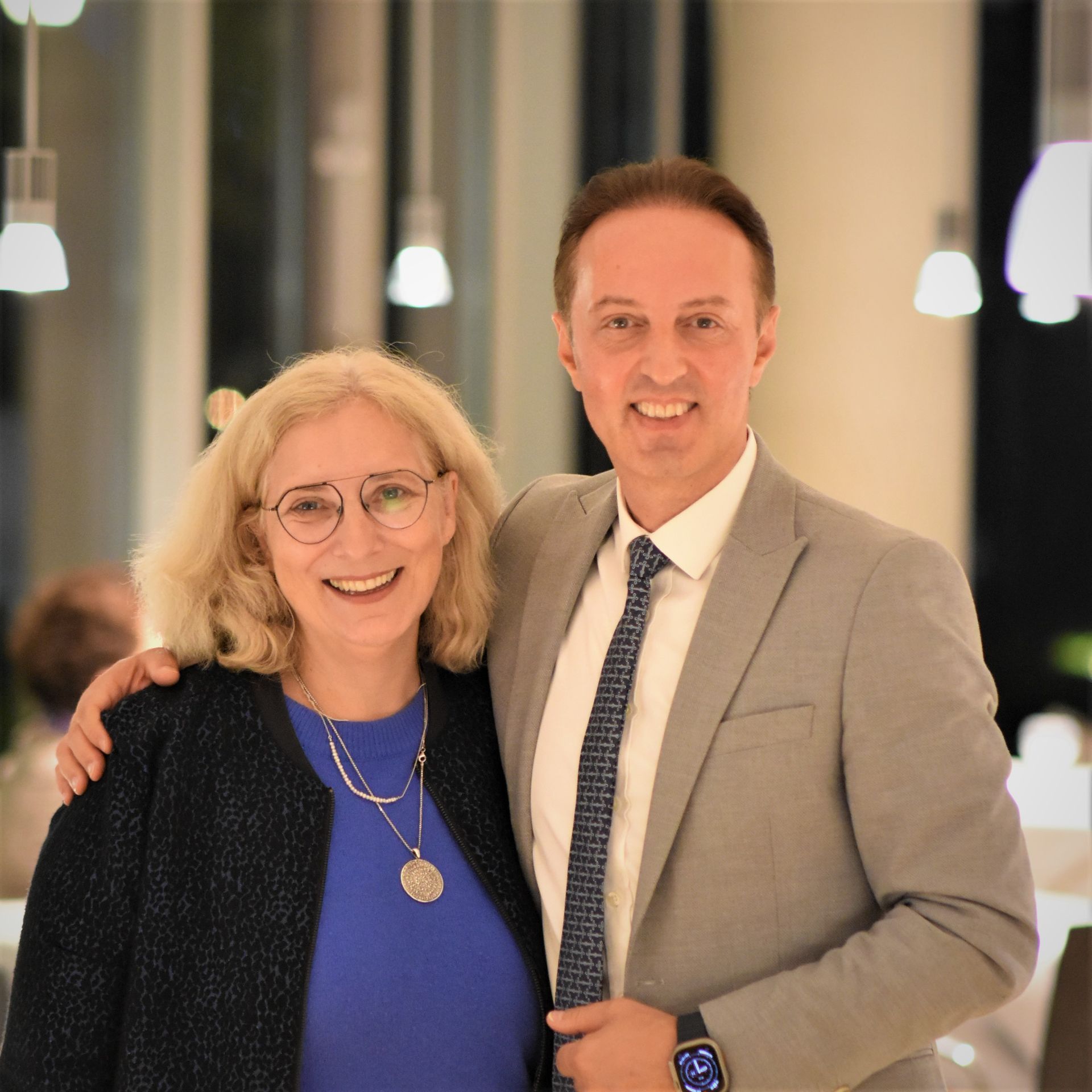 Eine Frau und ein Mann posieren für ein Foto. Sie befinden sich in einem Innenraum, lächeln und tragen formelle Kleidung.