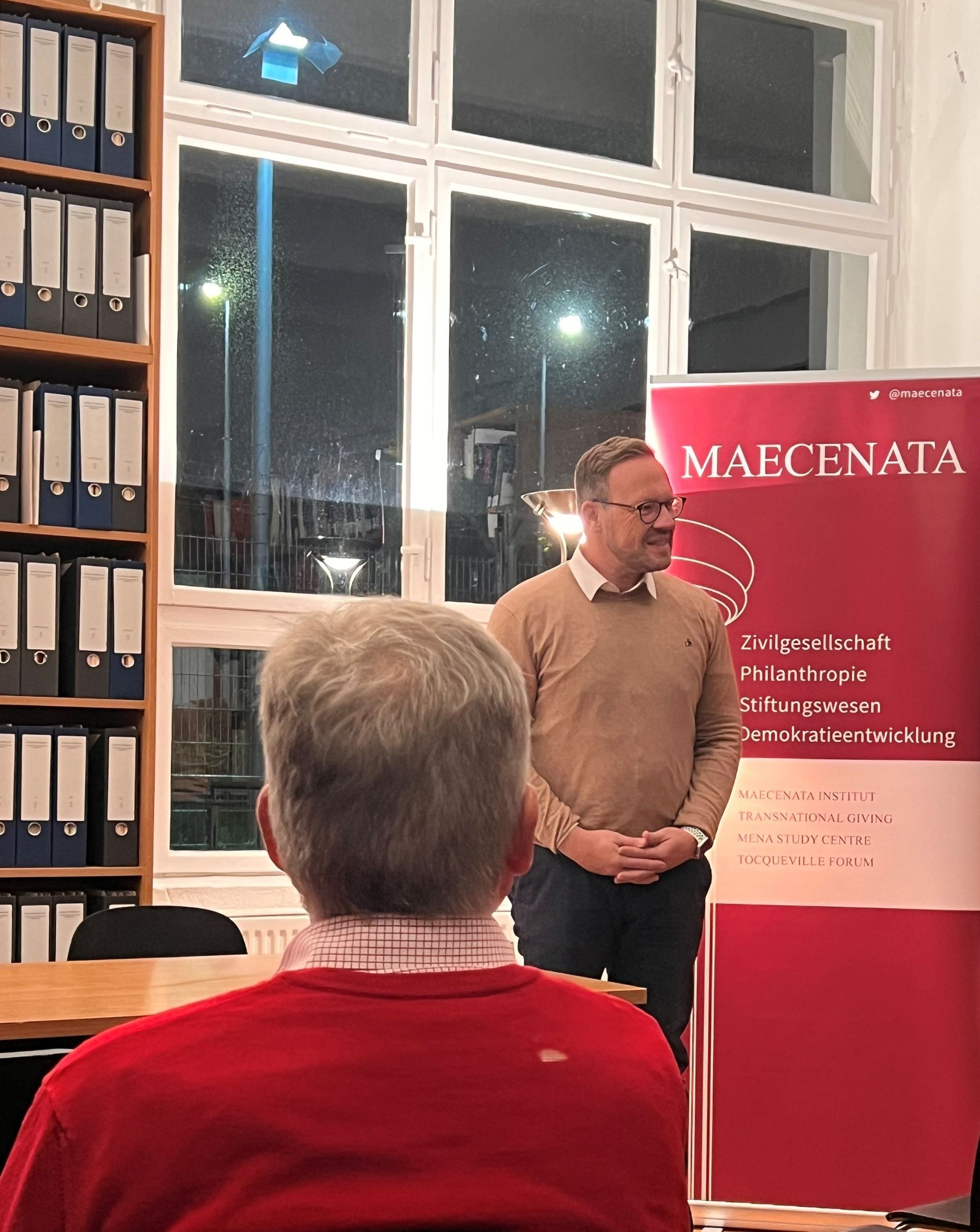 Ein Mann spricht bei einer Veranstaltung und steht neben einem Banner mit der Aufschrift „Maecenata“. Im Vordergrund ist eine Person in einem roten Pullover zu sehen.