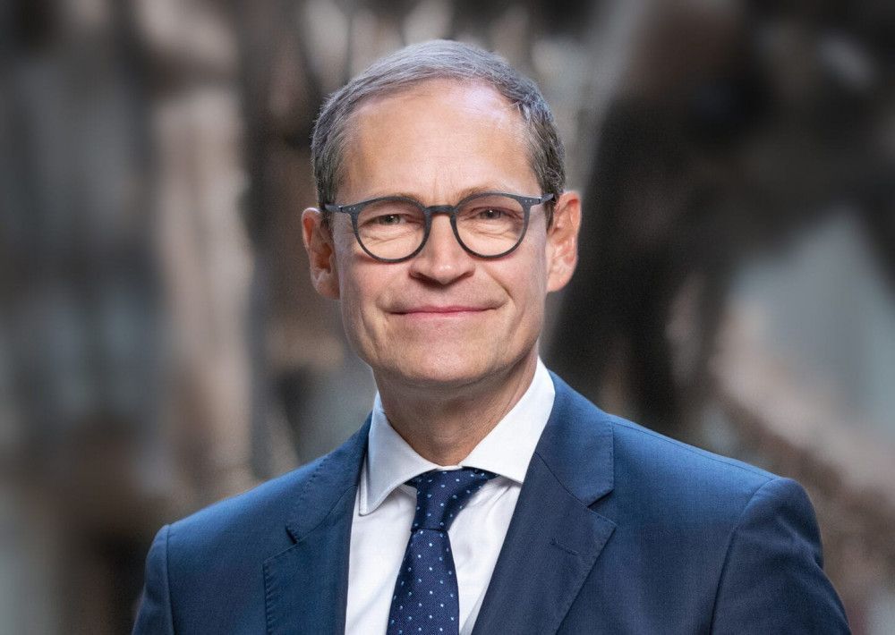 Mann im blauen Anzug und mit Brille lächelt in einem Innenraum.