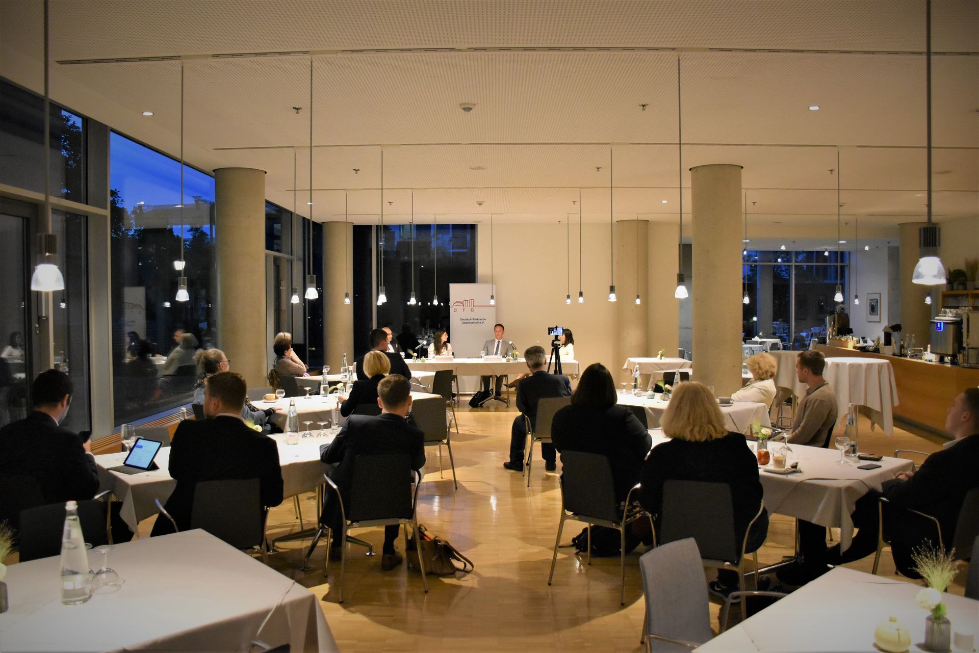 Menschen sitzen an Tischen in einem Raum und verfolgen eine Podiumsdiskussion bei sanfter Beleuchtung.